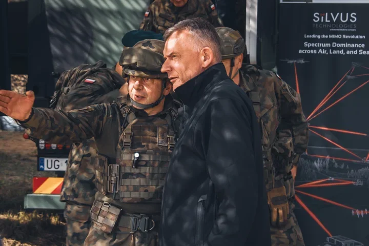 Żołnierz w kamuflażu gestykuluje podczas rozmowy z mężczyzną w czarnej kurtce przed pojazdem z napisem Silvus Technologies.