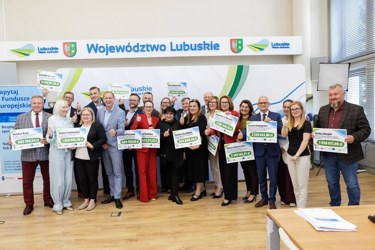 Grupa osób stoi na tle baneru Województwa Lubuskiego i trzyma tablice z kwotami dofinansowania dla gmin i miast.