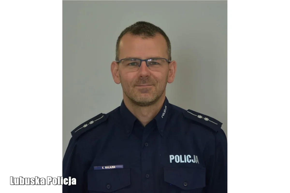 Mężczyzna w granatowym mundurze policyjnym z napisem „POLICJA” pozuje do zdjęcia portretowego na szarym tle.