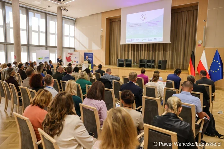 Uczestnicy konferencji siedzą w sali audytoryjnej, słuchając prelegenta przy podium, z flagami Niemiec, Polski i UE na scenie.