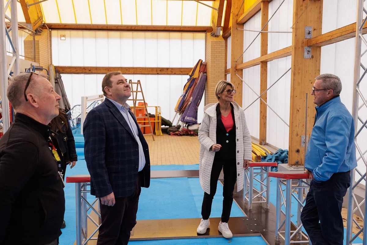 Cztery osoby rozmawiają we wnętrzu hali sportowej z drewnianą konstrukcją i niebieską podłogą.