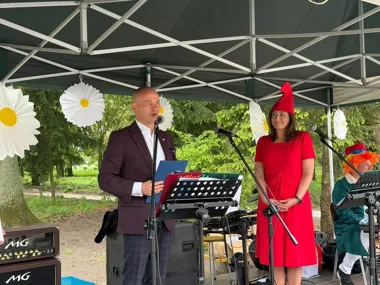 Mężczyzna w bordowej marynarce przemawia do mikrofonu na scenie pod namiotem, obok stoi kobieta w czerwonej sukience i czerwonym spiczastym czepku, a w tle sied