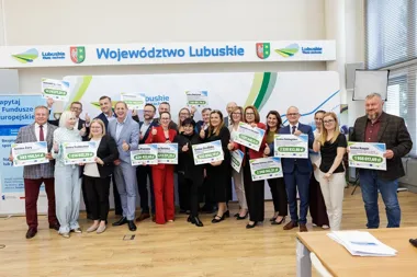 Grupa osób stoi na tle baneru Województwa Lubuskiego i trzyma tablice z kwotami dofinansowania dla gmin i miast.