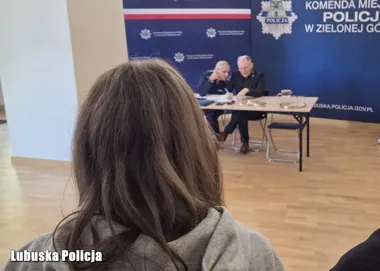 Kobieta siedzi na sali podczas spotkania z przedstawicielami policji przy stole przed ścianką z napisem Komenda Miejska Policji w Zielonej Górze.