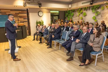 Mężczyzna w garniturze przemawia do zgromadzonych słuchaczy podczas konferencji w sali z drewnianą podłogą i zieloną ścianą.