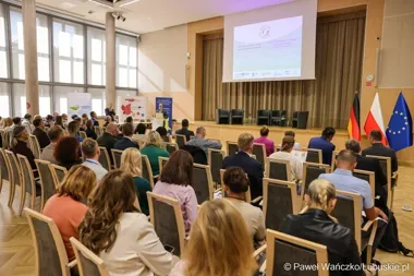 Uczestnicy konferencji siedzą w sali audytoryjnej, słuchając prelegenta przy podium, z flagami Niemiec, Polski i UE na scenie.