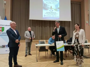 Mężczyzna w garniturze przemawia do mikrofonu na scenie sali konferencyjnej, trzymając torbę, w otoczeniu trzech innych osób; w tle ekran z prezentacją o trzeźw