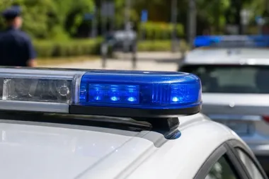 Zbliżenie na niebieski sygnał świetlny na dachu policyjnego radiowozu, w tle drugi radiowóz i stojący funkcjonariusz.