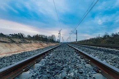 Nowo ułożony tor kolejowy z siecią trakcyjną biegnie przez przekop pod błękitnym niebem.
