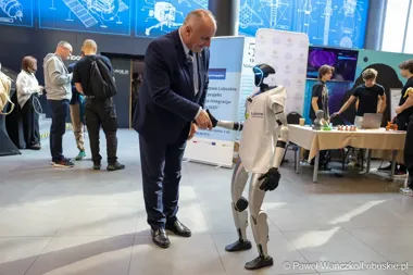 Mężczyzna w garniturze wita się z humanoidalnym robotem podczas wydarzenia w nowoczesnym centrum konferencyjnym.