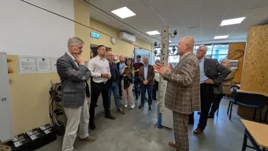 Mężczyzna w kraciastym garniturze gestykuluje do grupy osób podczas prezentacji lub wykładu w pomieszczeniu przypominającym warsztat lub laboratorium z urządzen