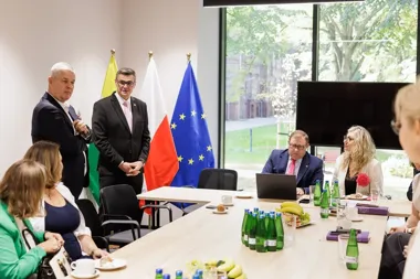 Spotkanie biznesowe w sali konferencyjnej z udziałem osób w eleganckich strojach, przy stole z napojami i przekąskami, w tle flagi Polski, Unii Europejskiej ora