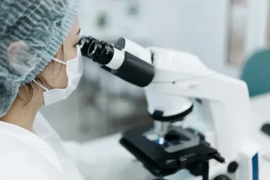 Kobieta w maseczce ochronnej i czepku wykonuje badania przy użyciu mikroskopu w laboratorium.