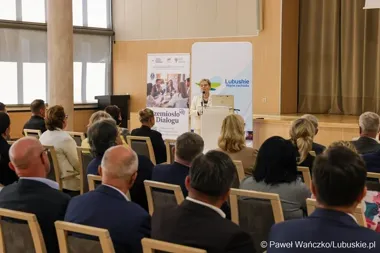 Kobieta w białym garniturze przemawia z mównicy do publiczności zgromadzonej w sali konferencyjnej, w tle banery z napisami „Lubuskie Warte zachodu” i „Rzemiosł