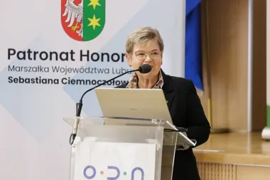 Kobieta w czarnej marynarce przemawia zza przezroczystego pulpitu z laptopem, na tle baneru z herbem i napisem „Patronat Honorowy Marszałka Województwa Lubuskie