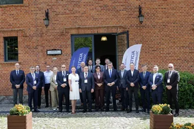 Grupa osób w eleganckim stroju stoi przed ceglanym budynkiem centrum konferencyjnego podczas wydarzenia z flagami reklamowymi.