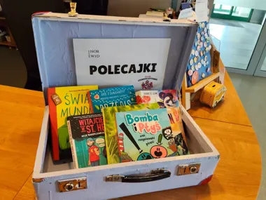 W otwartej, jasnoniebieskiej walizce na drewnianym stole leżą książki dla dzieci oraz kartka z napisem „POLECAJKI KLUB CICHEGO CZYTANIA”.