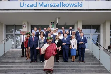 Grupa osób w eleganckim stroju biznesowym pozuje na schodach przed budynkiem Urzędu Marszałkowskiego, a w pierwszym planie stoi mężczyzna przebrany za postać z