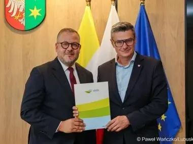 Dwóch mężczyzn w garniturach trzyma wspólnie dokument na tle flag.