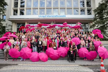 Grupa kobiet z różowymi parasolkami pozuje na schodach przed budynkiem Urzędu Marszałkowskiego.