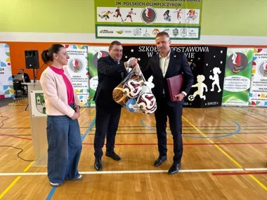 W sali gimnastycznej szkoły w Zielonej Górze dwaj mężczyźni wręczają sobie siatkę z piłkami, obok stoi kobieta, w tle banery szkolne i sponsorów.