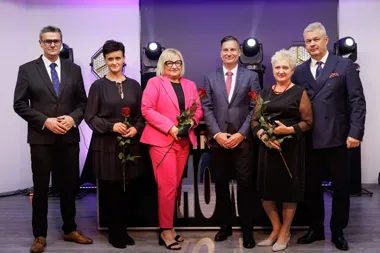 Sześć osób pozuje do grupowego zdjęcia na scenie podczas uroczystej gali, z czego trzy kobiety trzymają czerwone róże.
