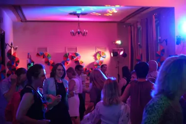 Goście bawią się na kolorowej imprezie w pomieszczeniu oświetlonym fioletowym i niebieskim światłem, z dekoracjami wiatraczków na ścianach.