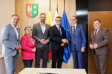 Sześć osób w eleganckich strojach biznesowych pozuje do zdjęcia w sali urzędowej, przy której dwoje mężczyzn ściska sobie dłonie, z herbem, flagą polską i flagą