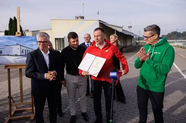 Grupa osób stoi na zewnątrz przed budynkiem sportowym podczas uroczystości, mężczyzna w czerwonej kurtce trzyma otwartą dokumentację i przemawia do mikrofonu TV