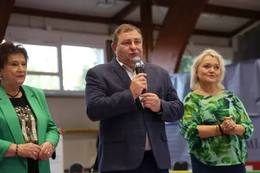 Mężczyzna w granatowej marynarce przemawia do mikrofonu w hali, stojąc między dwiema kobietami.