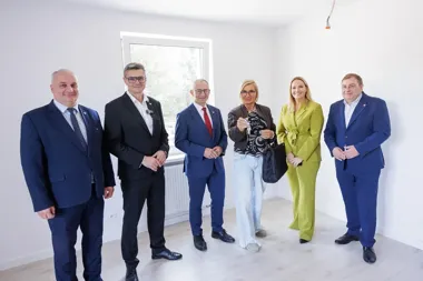Grupa sześciu osób, w tym kobieta trzymająca klucze, stoi w pustym, jasnym pomieszczeniu podczas uroczystego przekazania mieszkania.