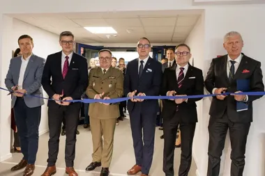 Grupa mężczyzn w garniturach i mundurze przecina niebieską wstęgę nożyczkami podczas uroczystego otwarcia w korytarzu biurowym.