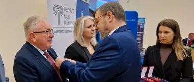 Mężczyzna w garniturze przypina odznaczenie innemu mężczyźnie podczas uroczystości w auli uniwersyteckiej, w tle baner Instytutu Nauk Prawnych Uniwersytetu Ziel