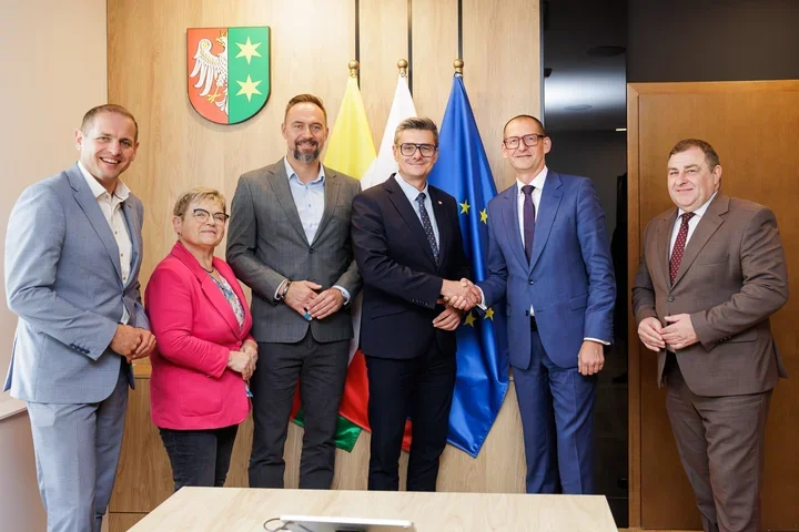 Sześć osób w eleganckich strojach biznesowych pozuje do zdjęcia w sali urzędowej, przy której dwoje mężczyzn ściska sobie dłonie, z herbem, flagą polską i flagą