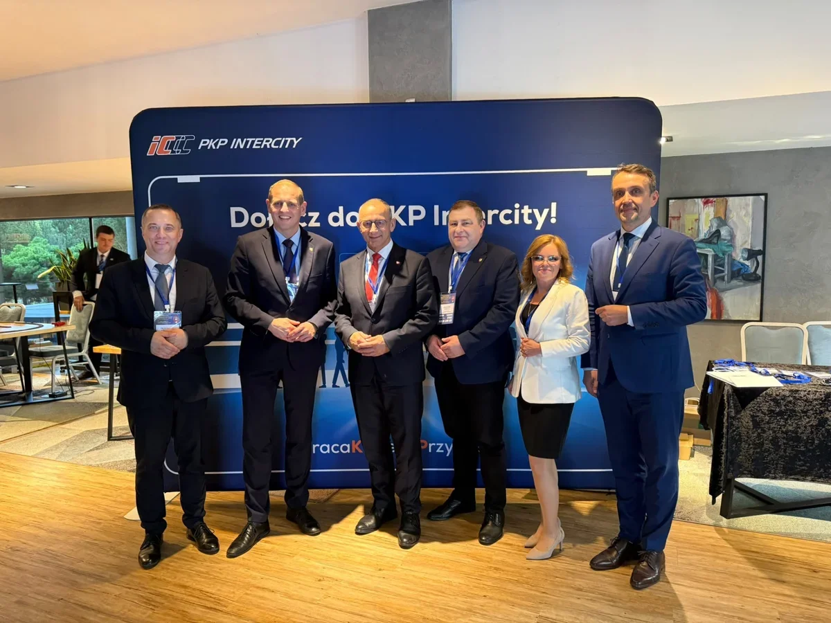 Grupa osób w strojach biznesowych pozuje do zdjęcia przed niebieską ścianką reklamową PKP Intercity podczas wydarzenia korporacyjnego.