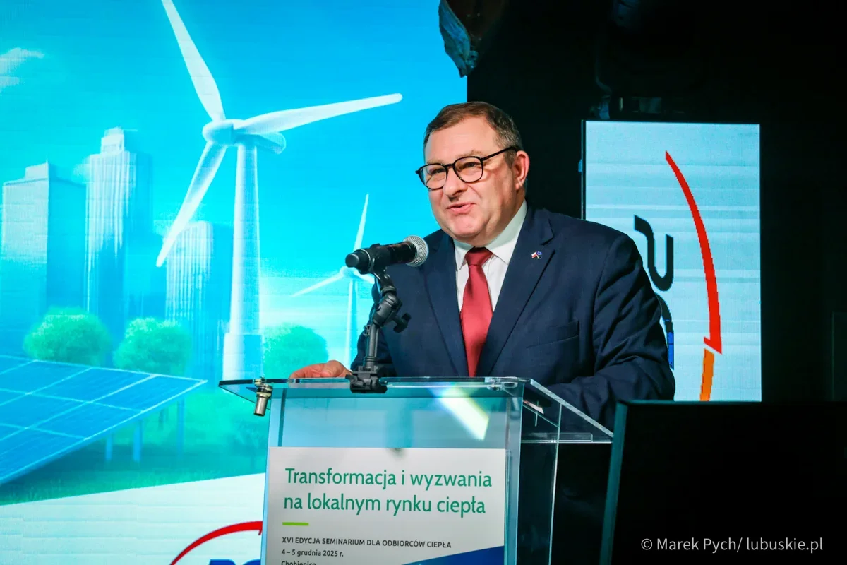 Mężczyzna w garniturze przemawia zza transparentnego pulpitu podczas seminarium o transformacji lokalnego rynku ciepła, w tle wizualizacje turbin wiatrowych i p