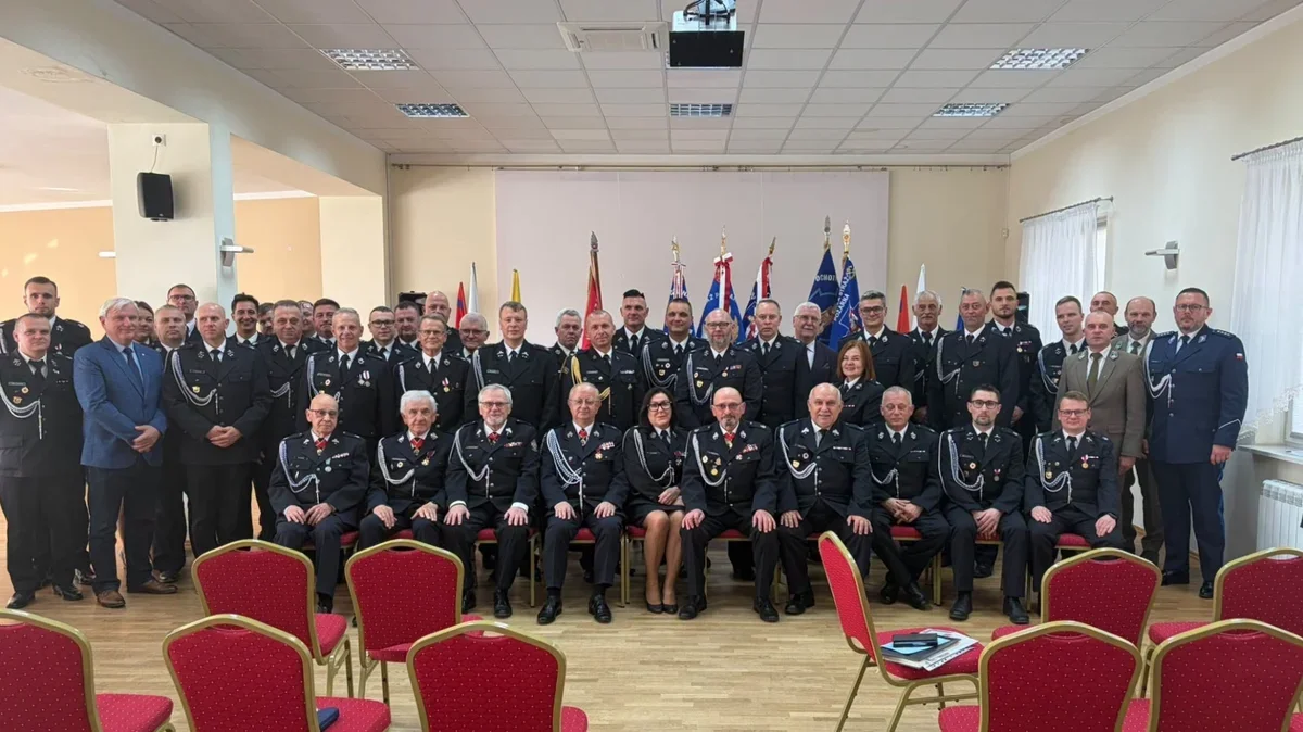Grupa mężczyzn i kilka kobiet, głównie w mundurach służb mundurowych, pozuje do pamiątkowego zdjęcia grupowego w sali konferencyjnej z flagami w tle.